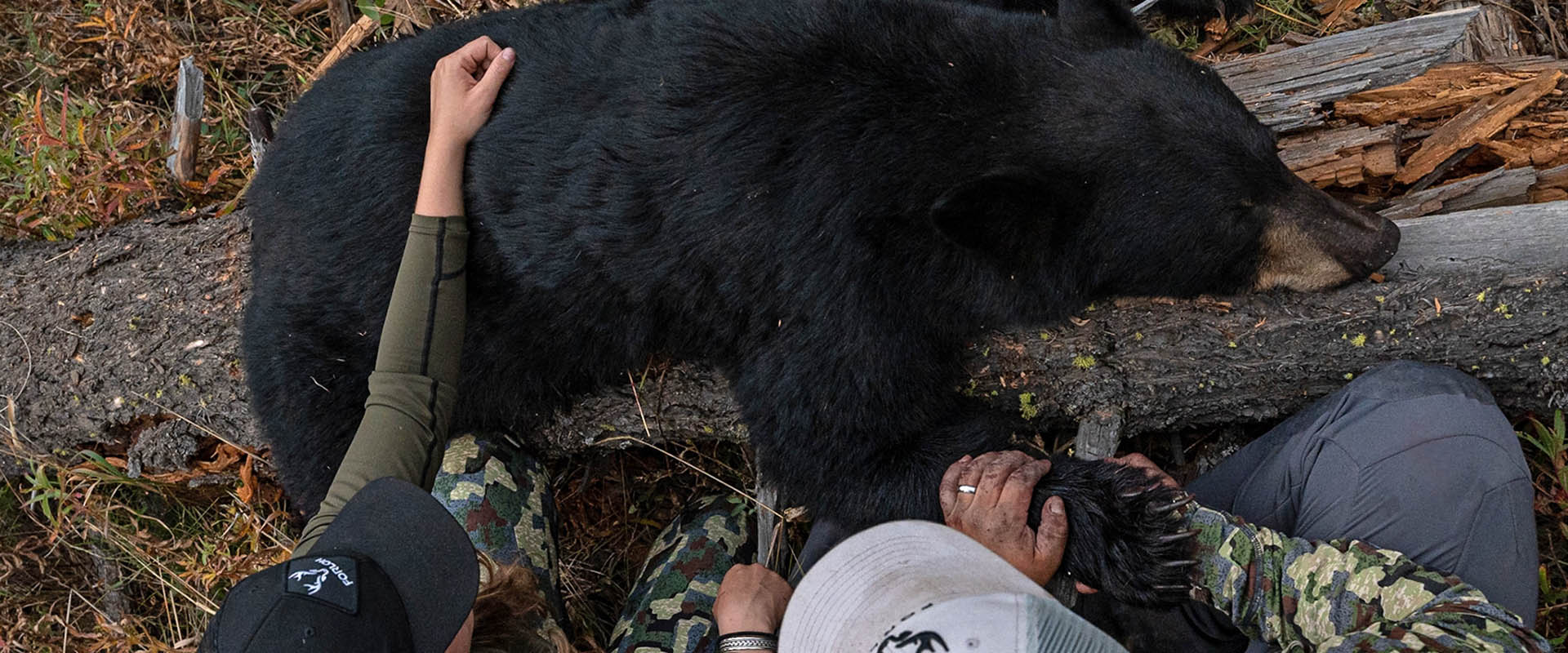 A woman and man hunter in camo gear sitting on the ground with with black bear