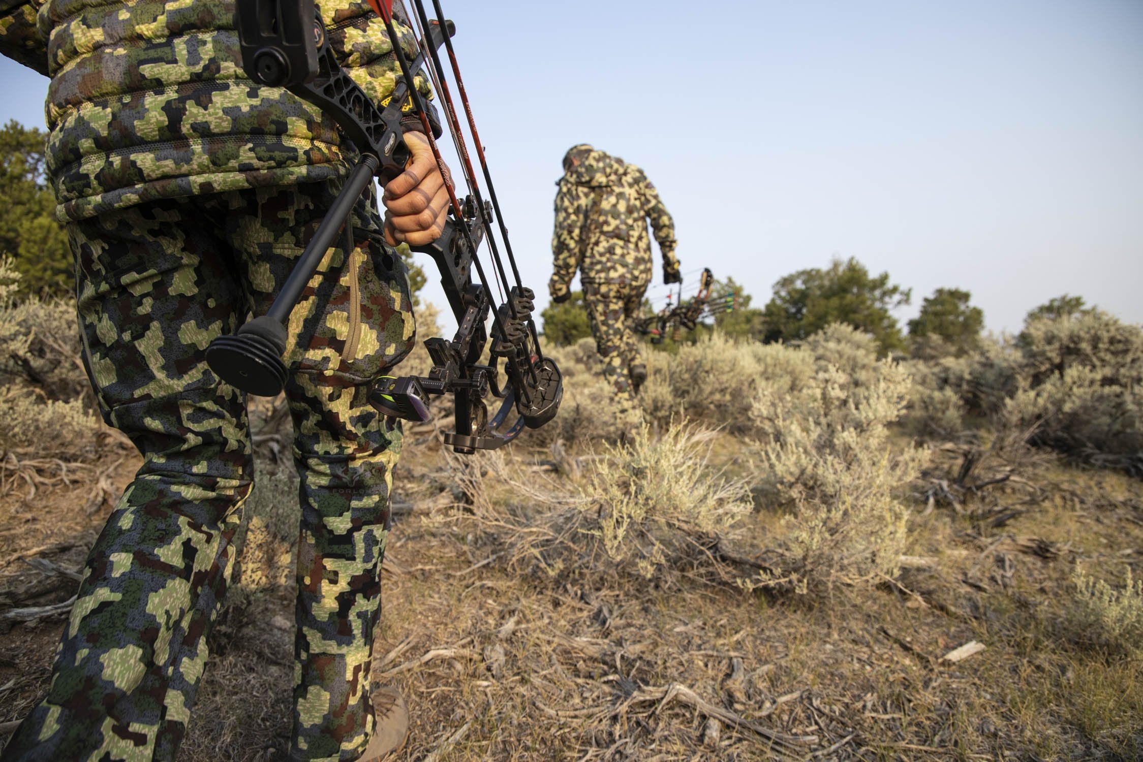 Hunter wearing deep cover camo walking with bow and arrow through terrain 